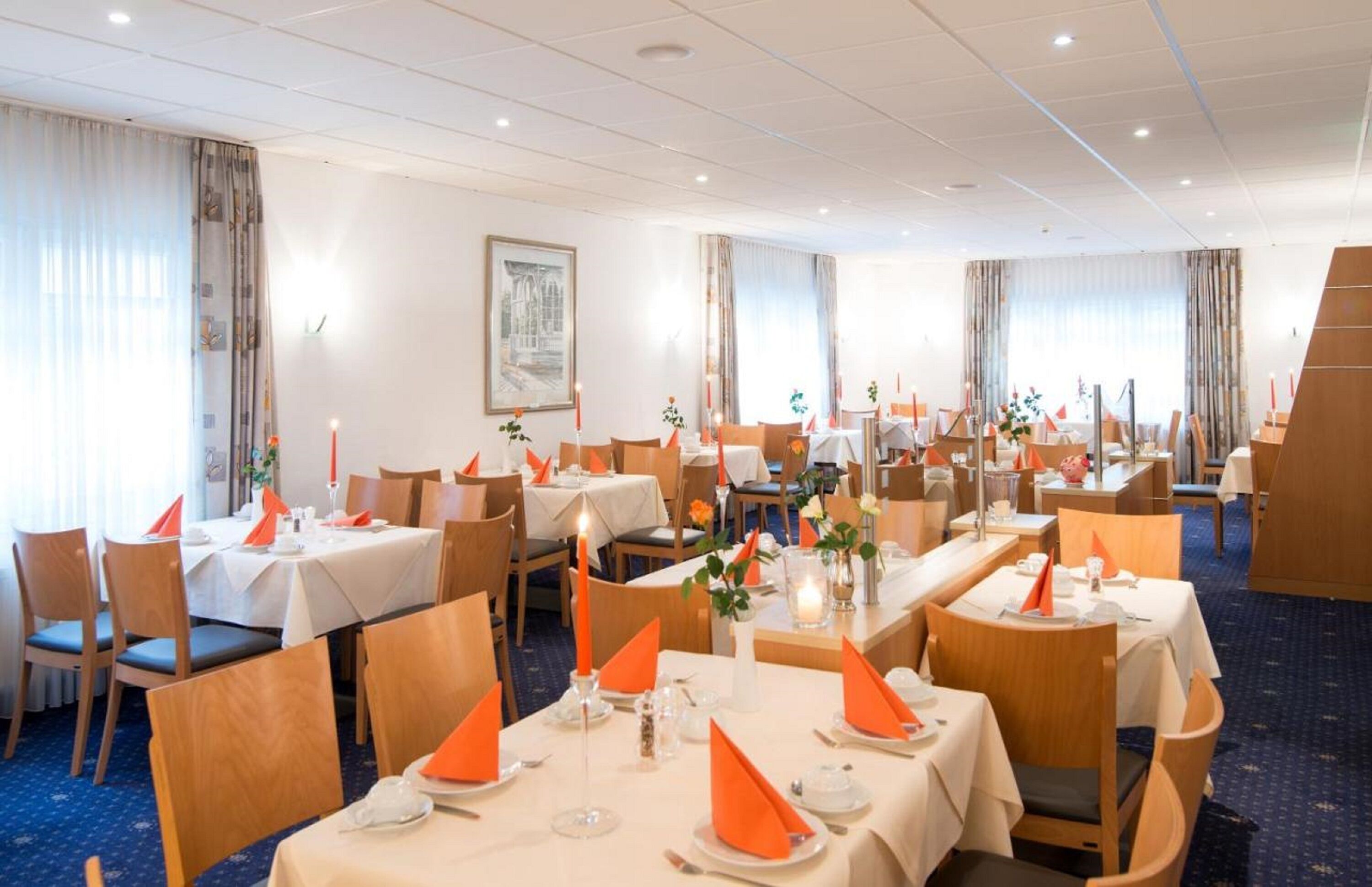 A hotel dining area with multiple tables set for dining, each with orange napkins and candles.