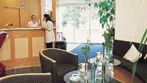A reception area with a counter, two receptionists, a seating area with a sofa and chairs, and a large window providing natural light.