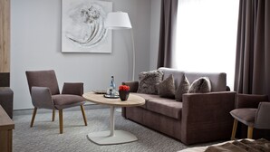 A modern hotel room with a grey sofa, two armchairs, a round wooden table, and a floor lamp. A large abstract painting adorns the wall, and a window with curtains provides natural light.