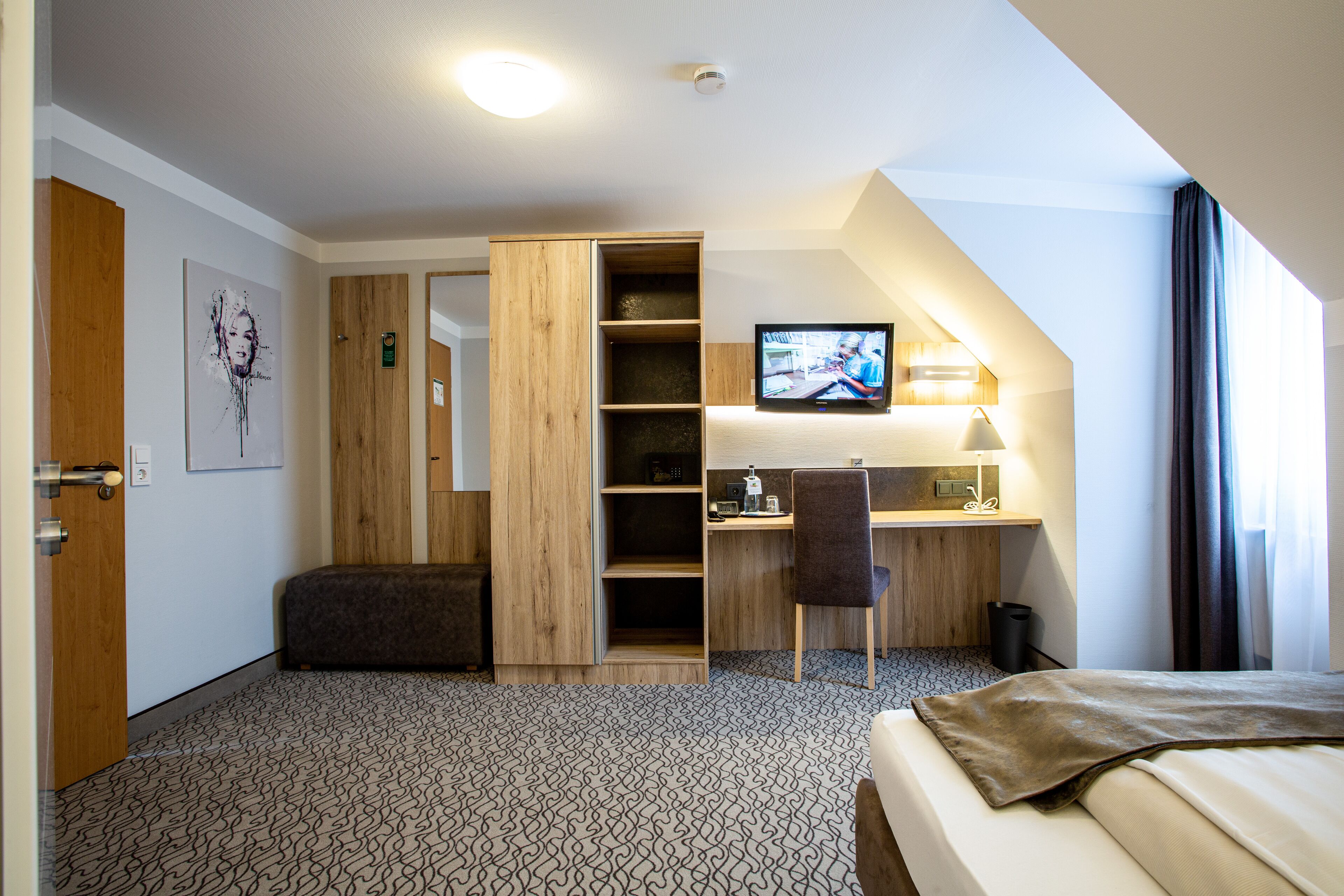 Habitación de hotel con cama, escritorio con silla, televisor, estantería y armario.