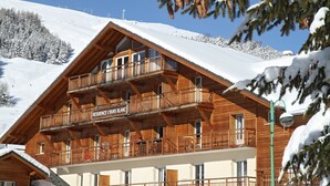 Un chalet en bois doté d’un balcon, une enseigne indiquant « RÉSIDENCE L’OURS BLANC », et une montagne enneigée en arrière-plan.