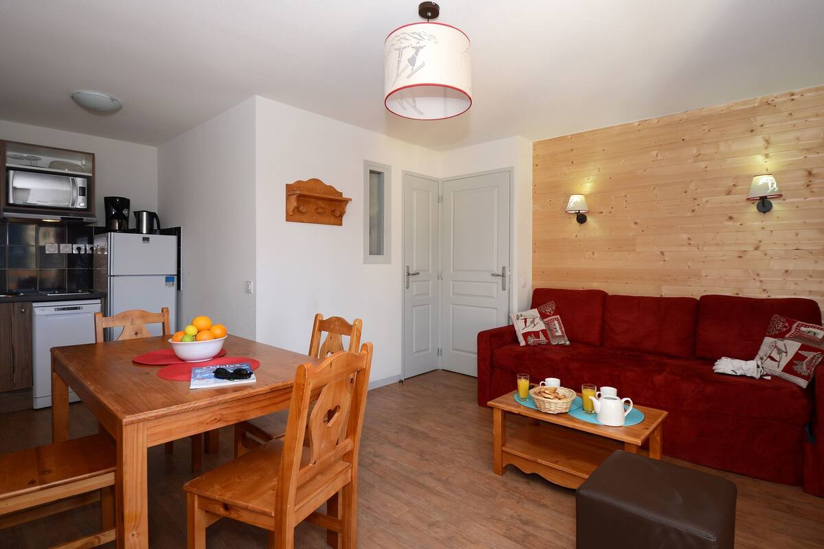 A compact living space with a wooden dining table, red sofa, and a kitchen area including a microwave and refrigerator.