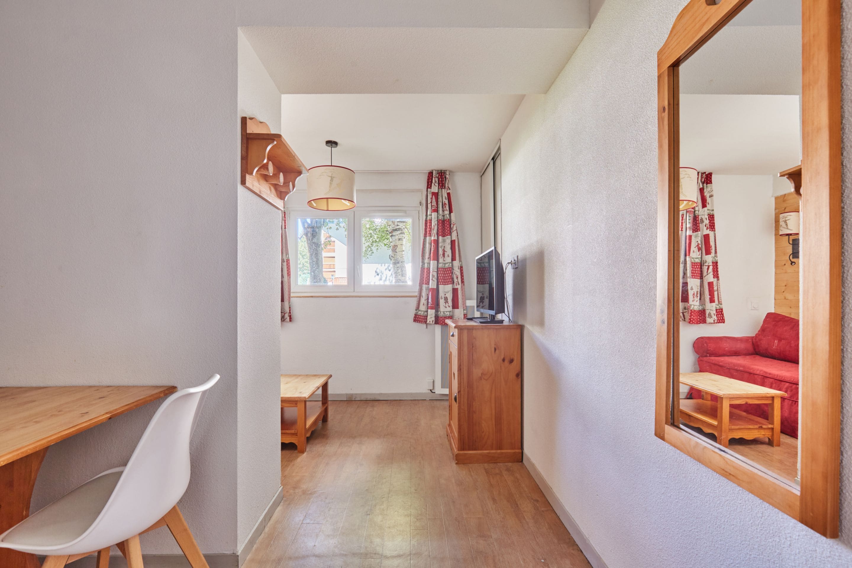 Un couloir étroit avec une table en bois, une chaise blanche, un téléviseur posé sur un meuble en bois et un grand miroir qui reflète un canapé rouge.