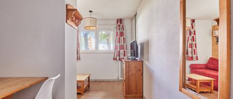 Un couloir étroit avec une table en bois, une chaise blanche, un téléviseur posé sur un meuble en bois et un grand miroir qui reflète un canapé rouge.