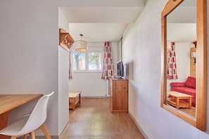 Un couloir étroit avec une table en bois, une chaise blanche, un téléviseur posé sur un meuble en bois et un grand miroir qui reflète un canapé rouge.
