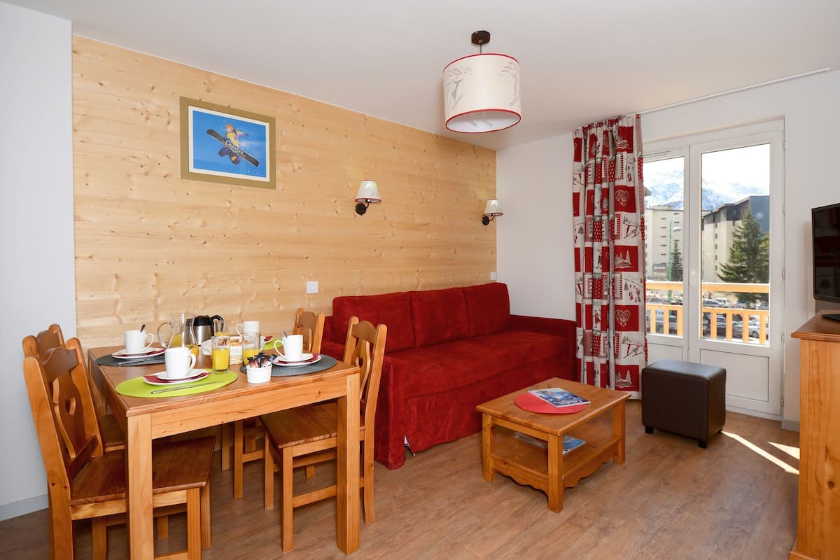 A cozy living room with a wooden dining table set for breakfast, a red sofa, a television, and a window with a view of buildings and mountains.