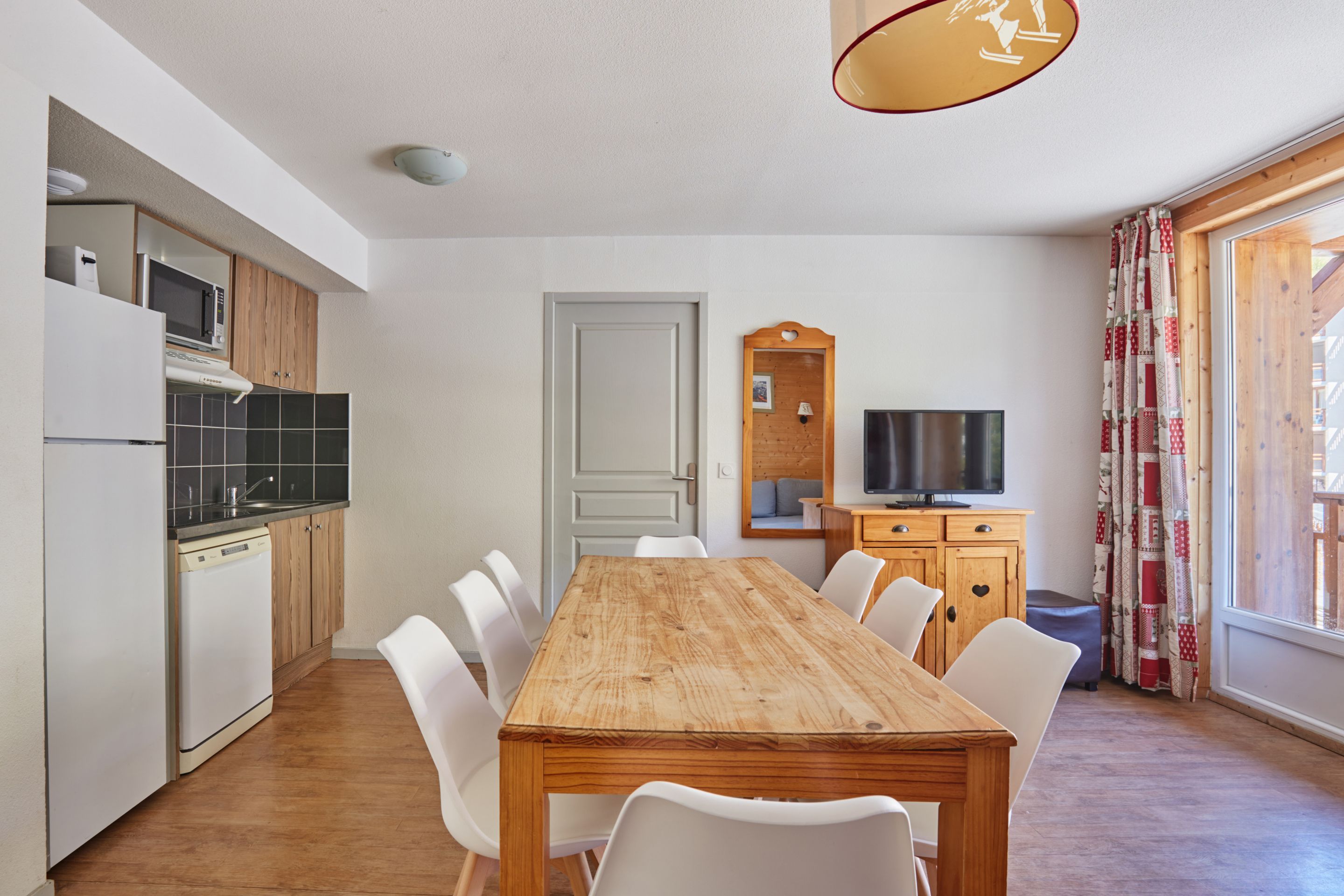 Eine kompakte Küche mit modernen Geräten, ein Essbereich mit einem Holztisch und weißen Stühlen sowie ein Fernseher auf einer Holzkommode.