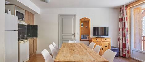 Eine kompakte Küche mit modernen Geräten, ein Essbereich mit einem Holztisch und weißen Stühlen sowie ein Fernseher auf einer Holzkommode.