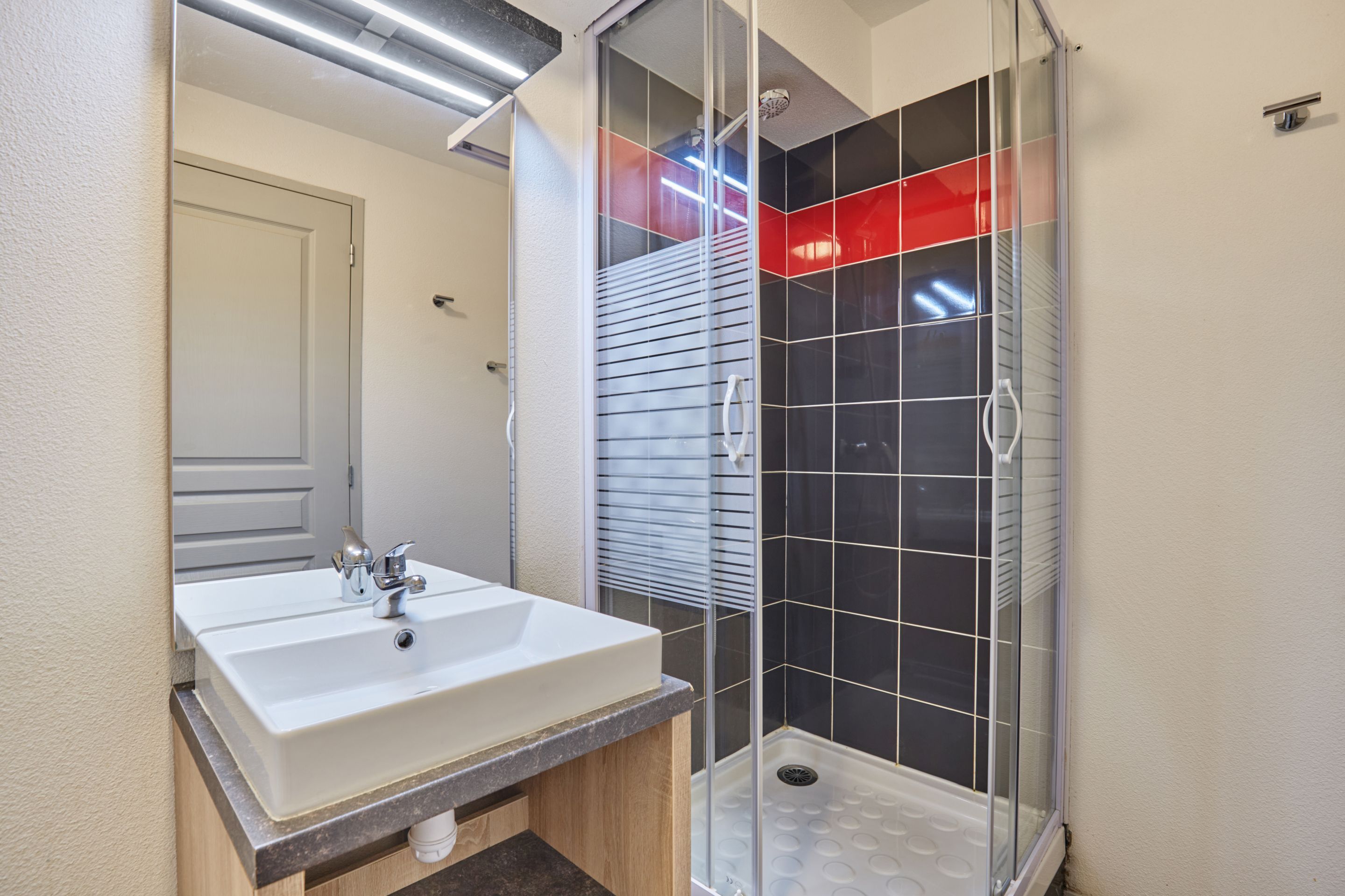 A bathroom with a white sink, a mirror, and a glass-enclosed shower with black tiles.