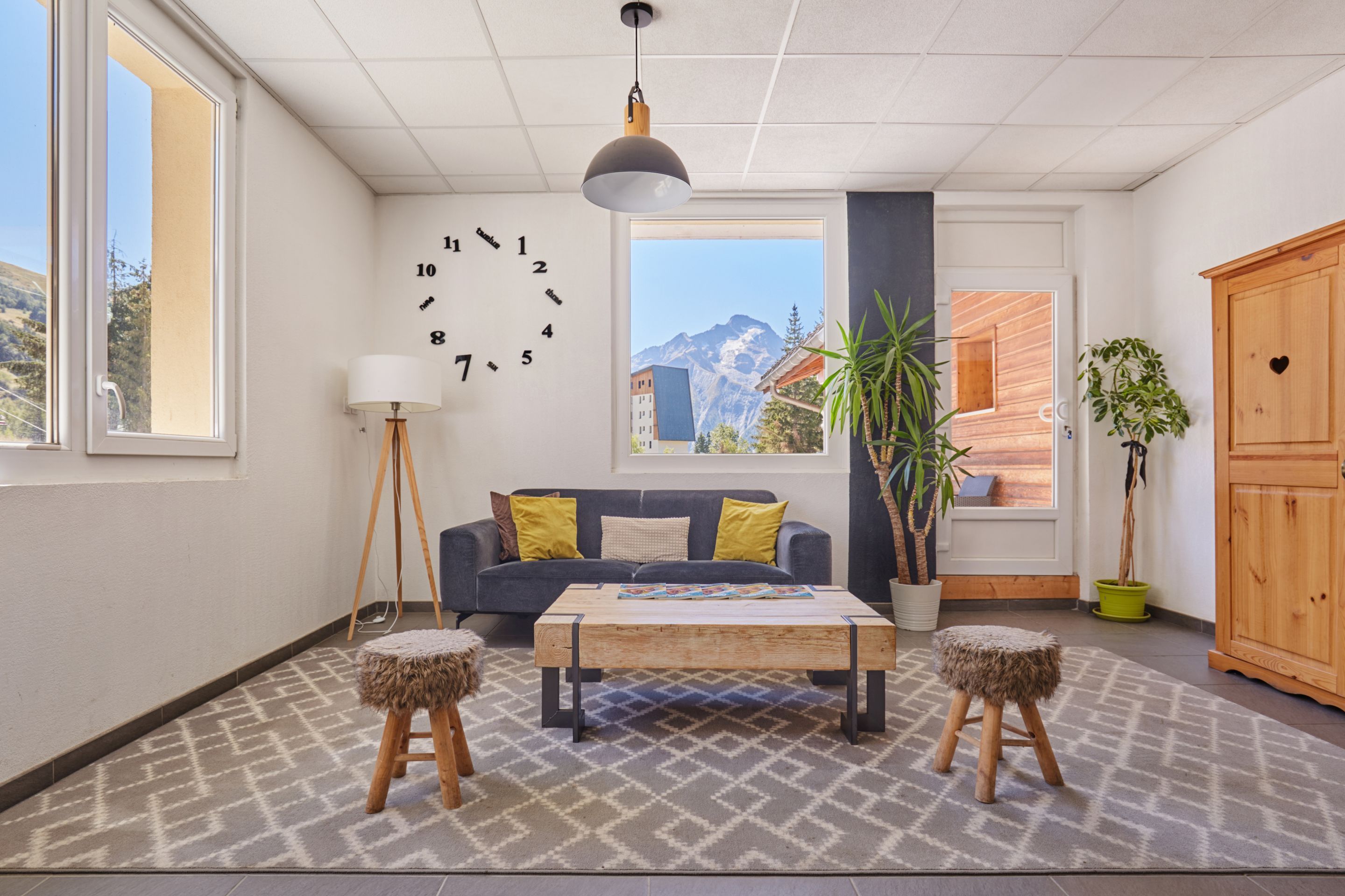 A modern living room with a sofa, coffee table, and two stools. There is a large window with a view of mountains, a clock on the wall, and a plant.