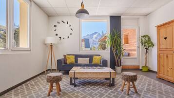A modern living room with a sofa, coffee table, and two stools. There is a large window with a view of mountains, a clock on the wall, and a plant.