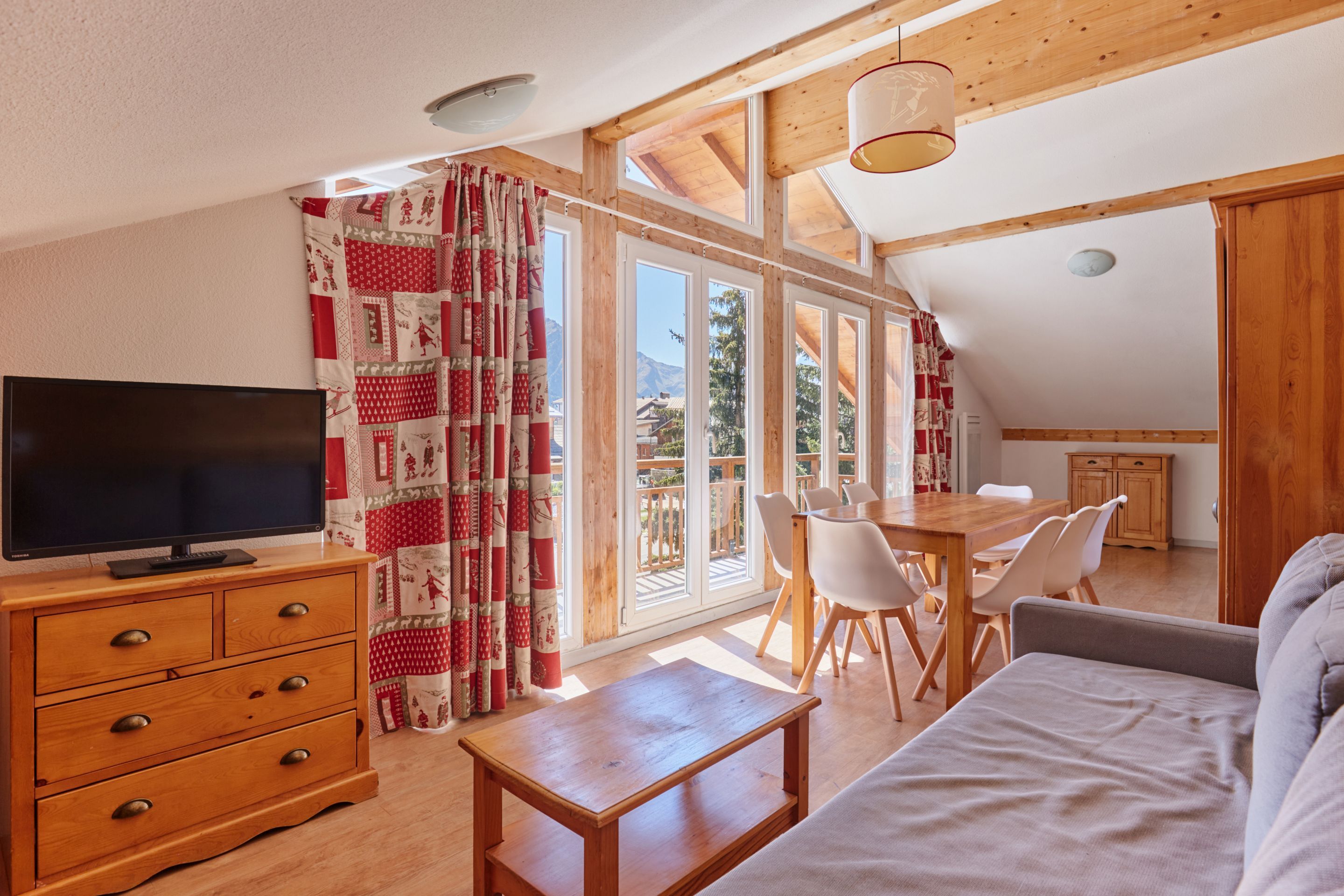 Un salon chaleureux avec un téléviseur, des meubles en bois et de grandes fenêtres donnant sur l’extérieur.