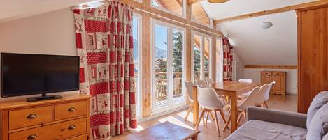 Un salon chaleureux avec un téléviseur, des meubles en bois et de grandes fenêtres donnant sur l’extérieur.