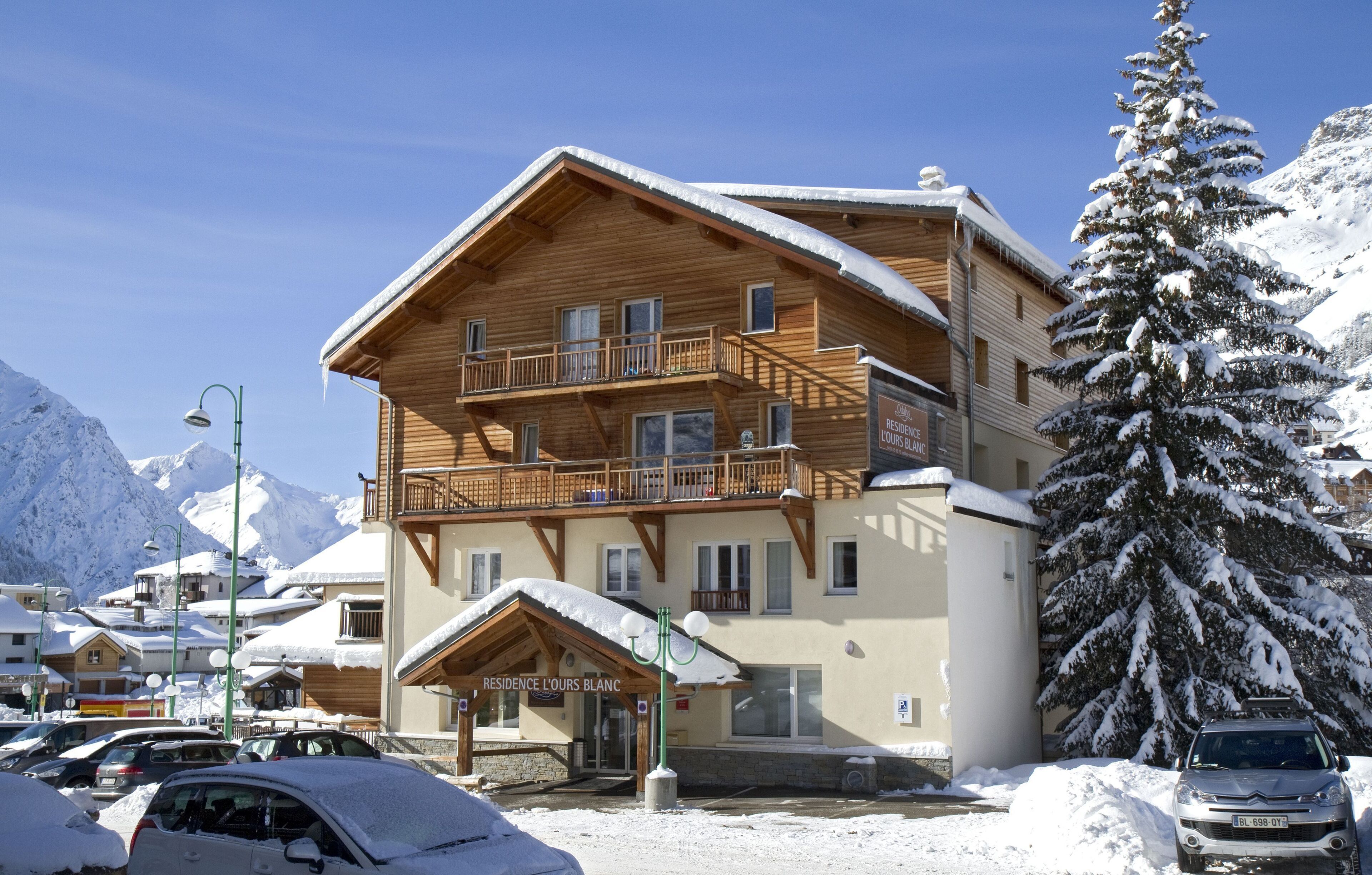 A multi-story wooden building with balconies, a sign reading "RESIDENCE L'OURS BLANC," and a snow-covered tree nearby.