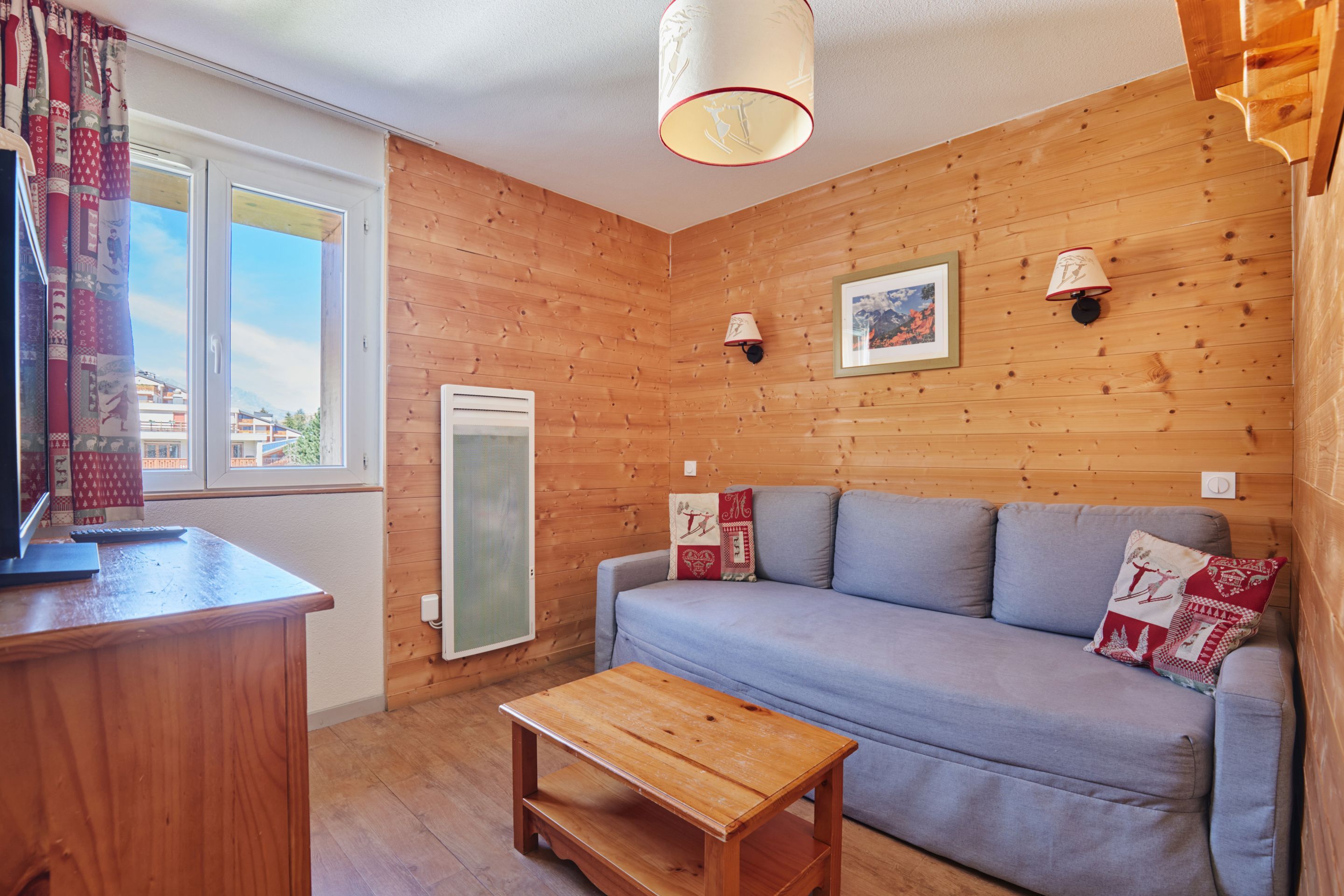 Una habitación acogedora con una pared de madera, un sofá gris, una pequeña mesa de madera y una ventana con cortinas.
