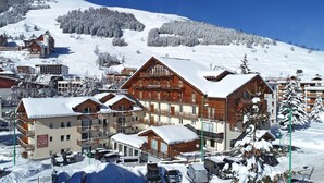 Une ville de montagne enneigée, avec des bâtiments en bois, dont un hôtel doté de balcons et d’une enseigne indiquant « Oberlies Residence L’Ours Blanc ».
