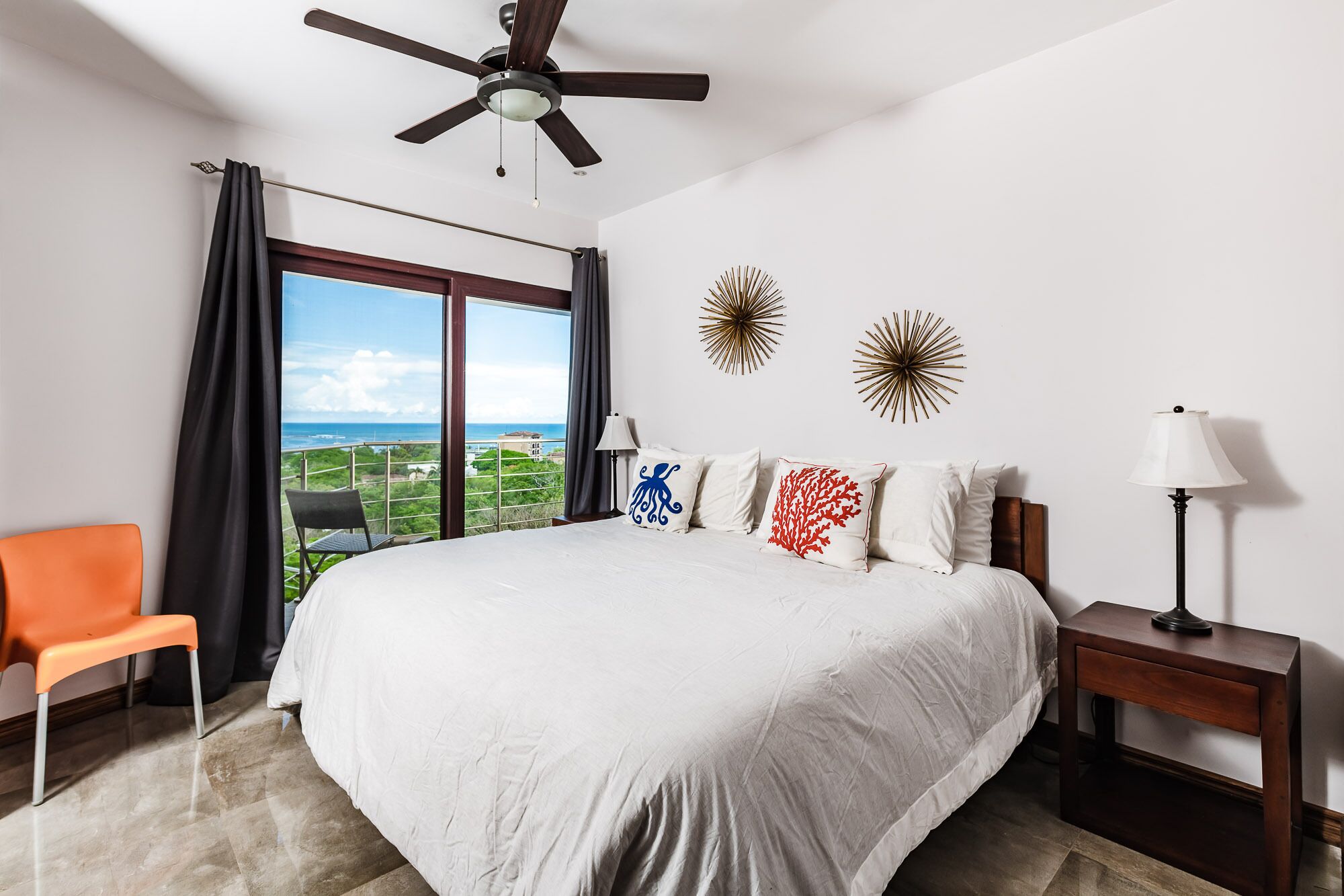 A bedroom with a large bed, a ceiling fan, a view of the ocean, and two wall decorations.
