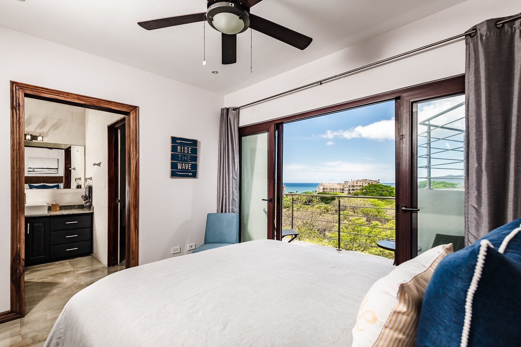 A bedroom with a bed, a ceiling fan, a bathroom with a mirror, and a balcony with a view of the sea.