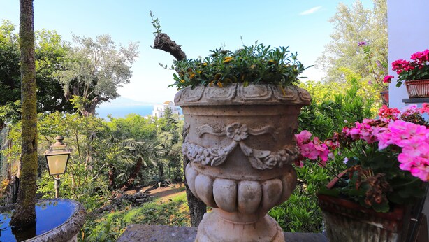 Puutarha, jossa on kivinen urnamainen koristeastia, lyhtypylväs ja näkymä merelle.