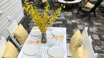 Une terrasse extérieure avec une table blanche dressée pour quatre personnes, entourée de chaises blanches dotées de coussins jaunes.
