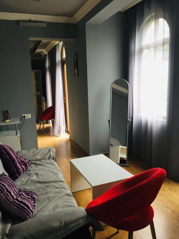 A modern living room with a grey sofa, a red chair, a white coffee table, and a floor lamp. There is a large window with curtains, a mirror on the wall, and a door leading to another room. - OLD CITY Boutique Hotel (Batumi)