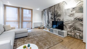 A modern living room with a large sectional sofa, a flat-screen TV on a white stand, a large window with a view of a building, and a tropical-themed mural on the wall. - Solemar Sicilia - Galleria Maqueda (Palermo)