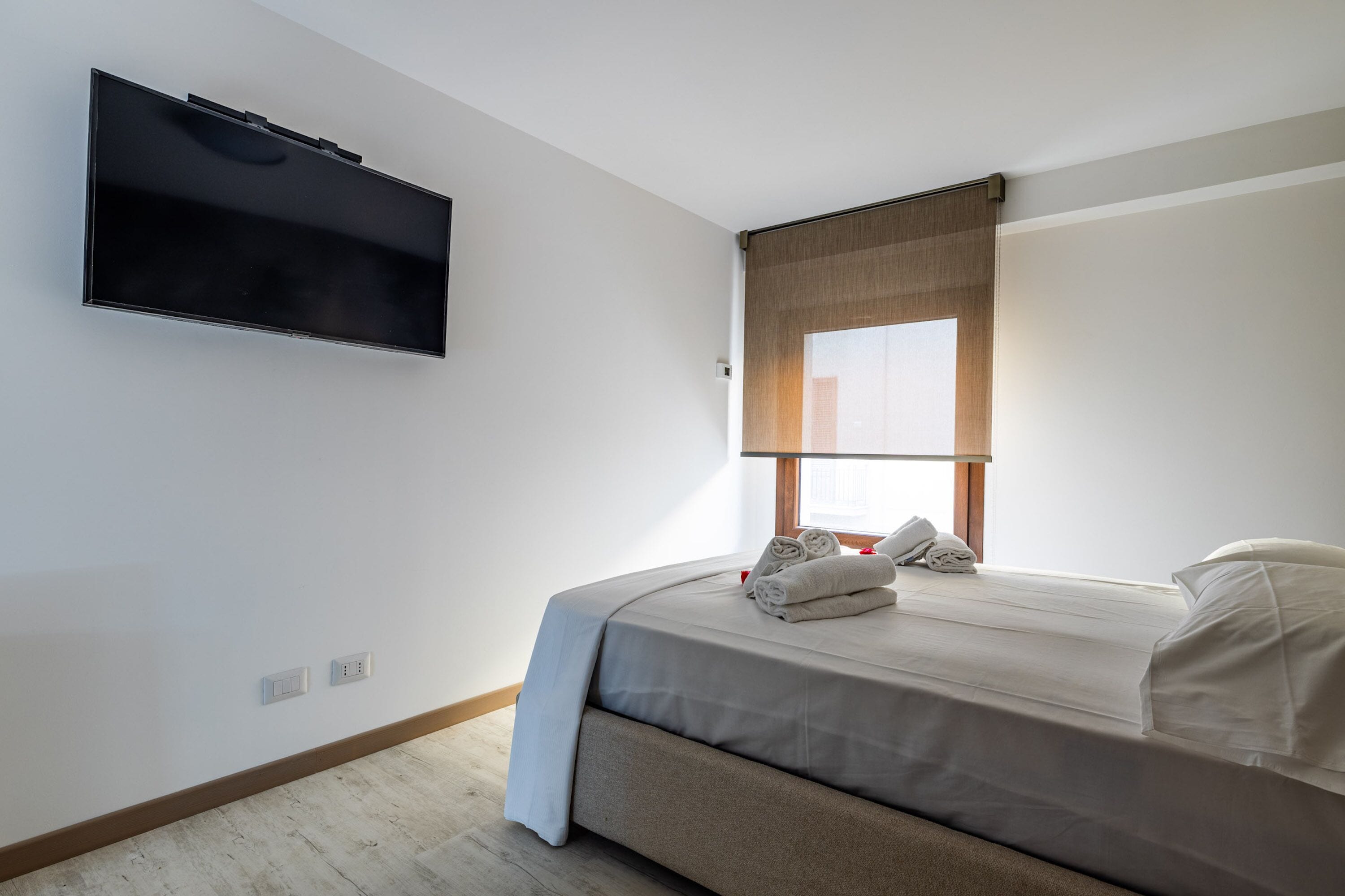 Een moderne hotelkamer met een flatscreen televisie aan de muur, een bed met een beigekleurige matras en witte lakens, en een raam met houten jaloezieën.