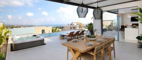 A modern outdoor dining area with a pool, wooden furniture, and a view of the sea.