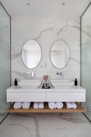 A modern bathroom with two sinks, marble walls, and a wooden shelf with towels. - Golden Shore (Pegeia)