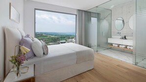 A modern bedroom with a large bed, a glass-enclosed bathroom, and a view of the countryside. - Golden Shore (Pegeia)