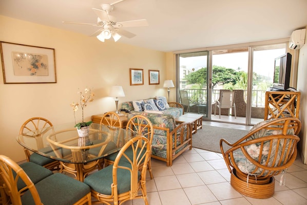 A living room with a glass dining table, wicker chairs, a sofa, and a ceiling fan. There is a balcony with a view of greenery.