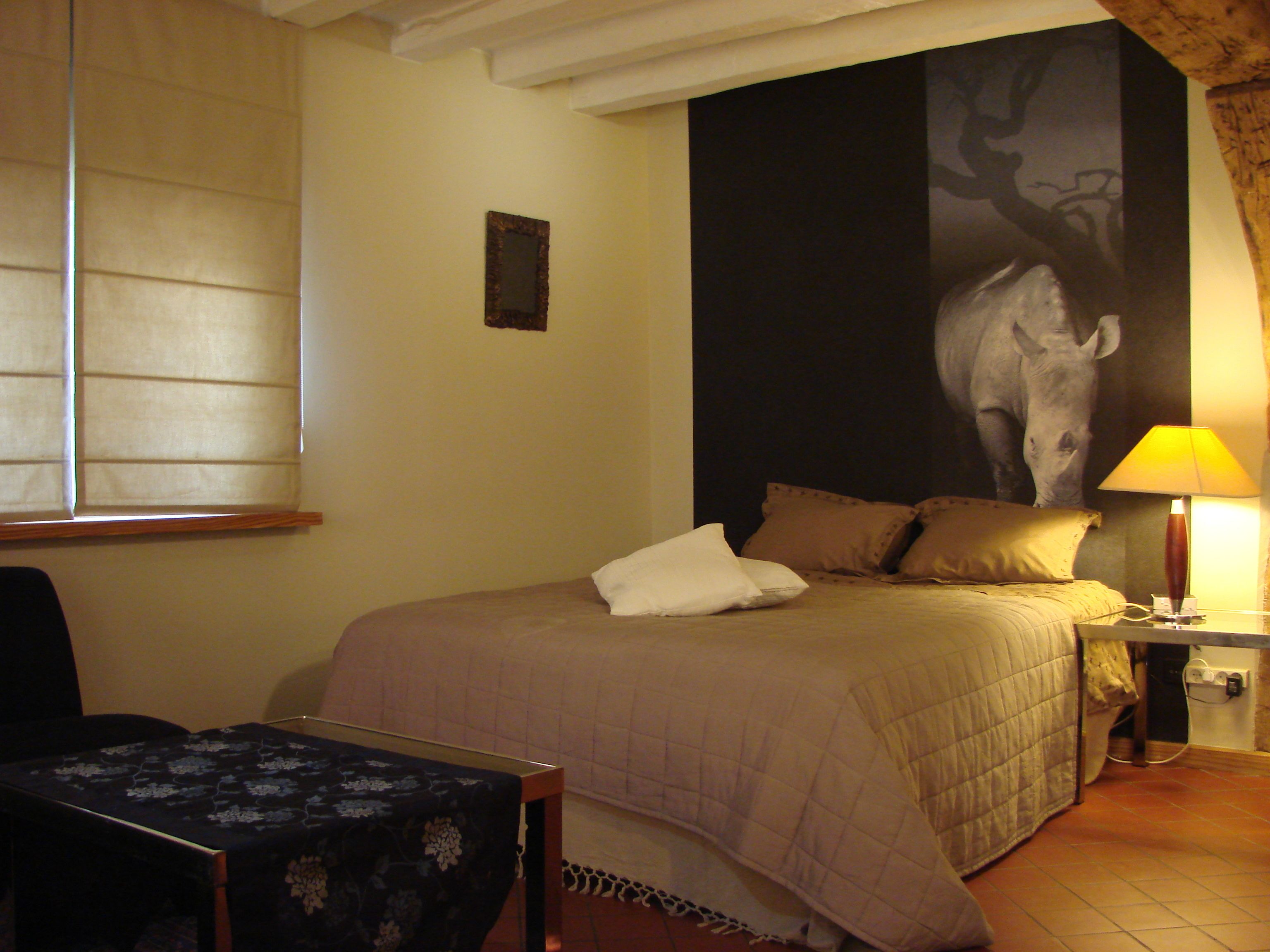 A hotel room with a bed, a nightstand with a lamp, a chair, and a large wall painting.