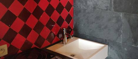 Una cocina con un lavamanos blanco, encimera negra, salpicadero a cuadros rojo y negro, y pared de piedra.