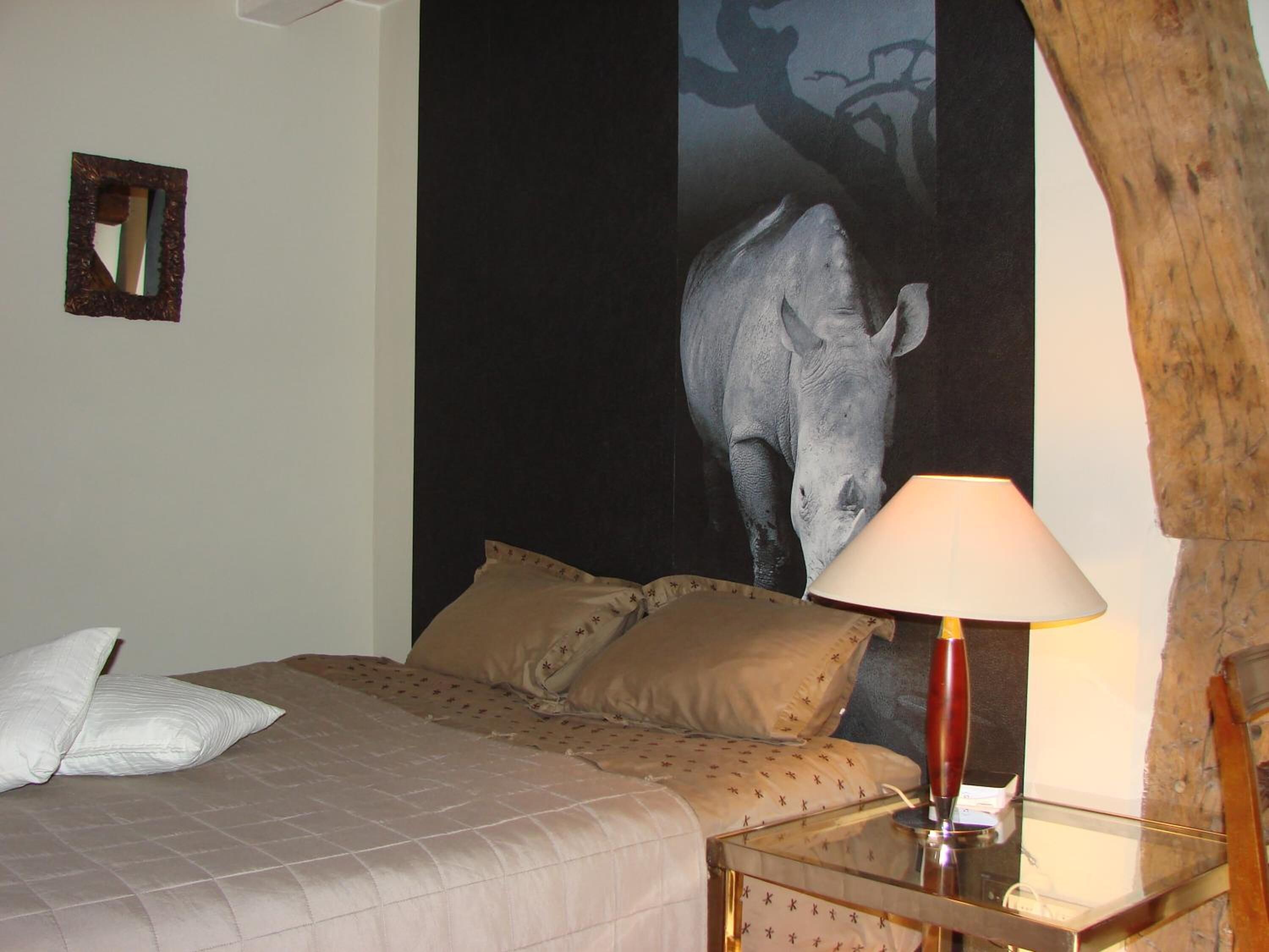 A bedroom with a bed, pillows, a nightstand, a lamp, and a large wall tapestry of a rhinoceros.