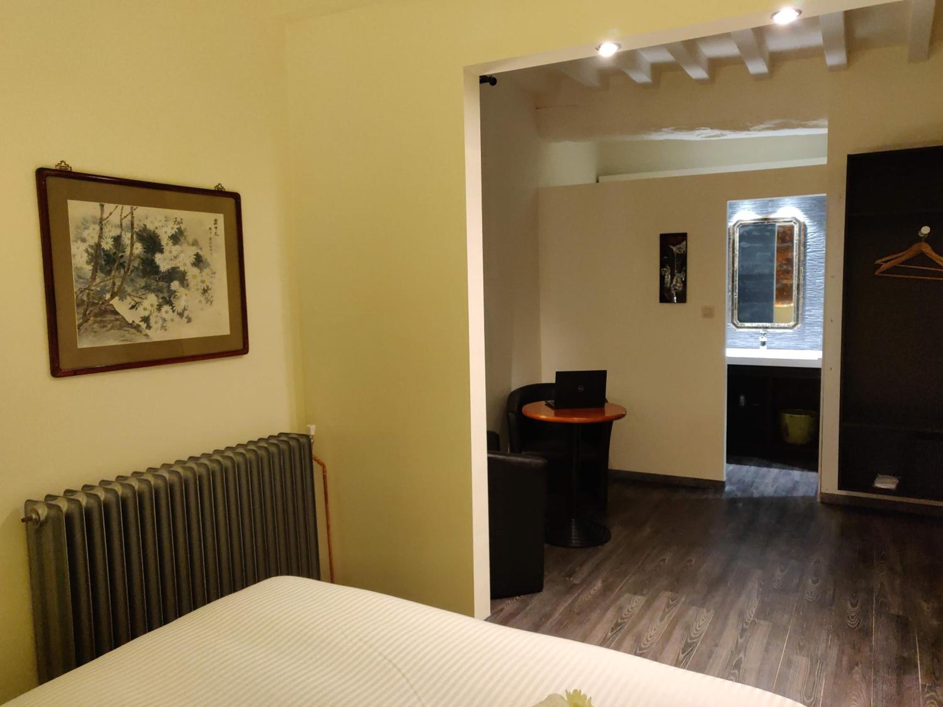 Quarto de hotel com cama, radiador, quadro emoldurado, pequena mesa com um laptop e espelho.