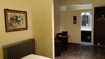 Quarto de hotel com cama, radiador, quadro emoldurado, pequena mesa com um laptop e espelho.
