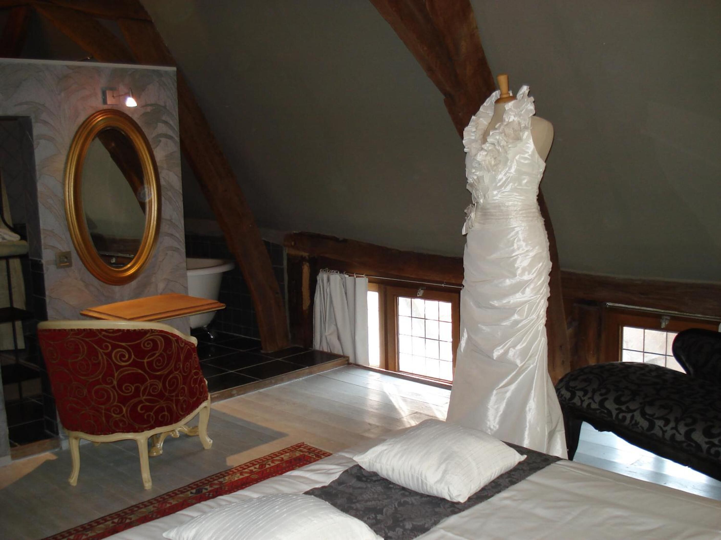 Une chambre à coucher avec un lit, une chaise rouge, une commode en bois, un miroir et une robe sur un mannequin.