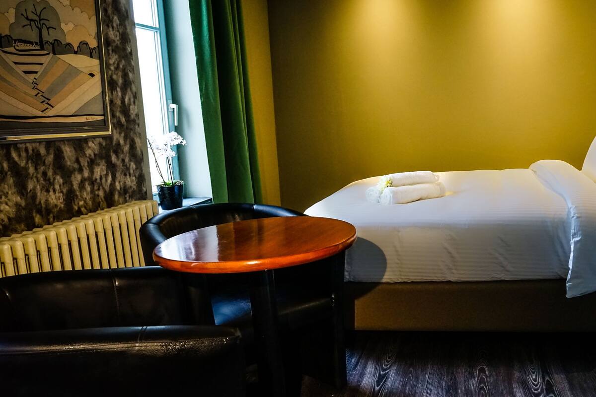 A hotel room with a bed, a chair, a small table, a radiator, and a painting on the wall.