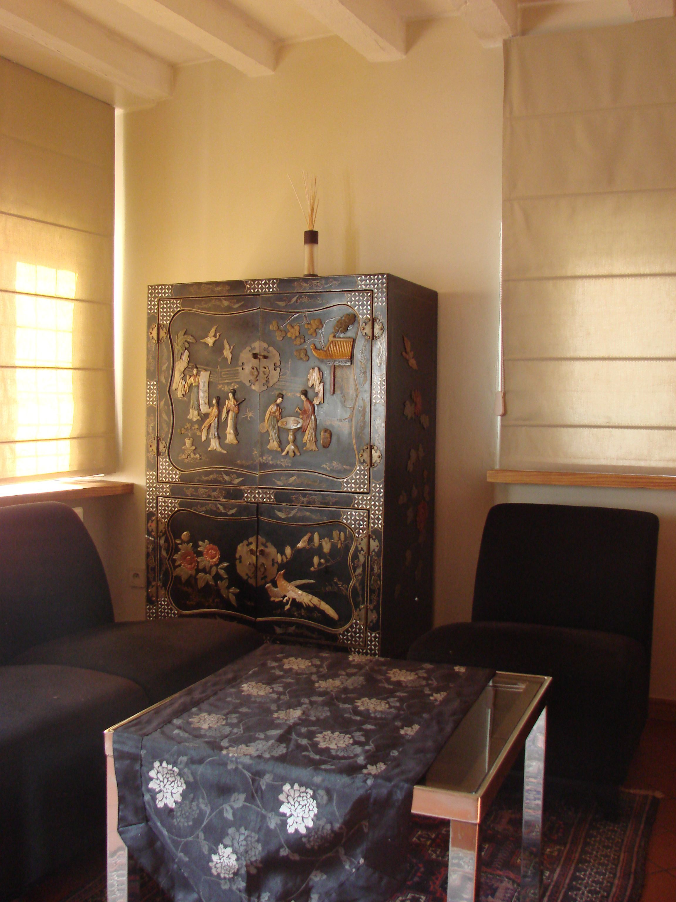 Een kamer met een klassiek kastje, een tafeltje met een bloemrijk tafelkleed, en twee stoelen.
