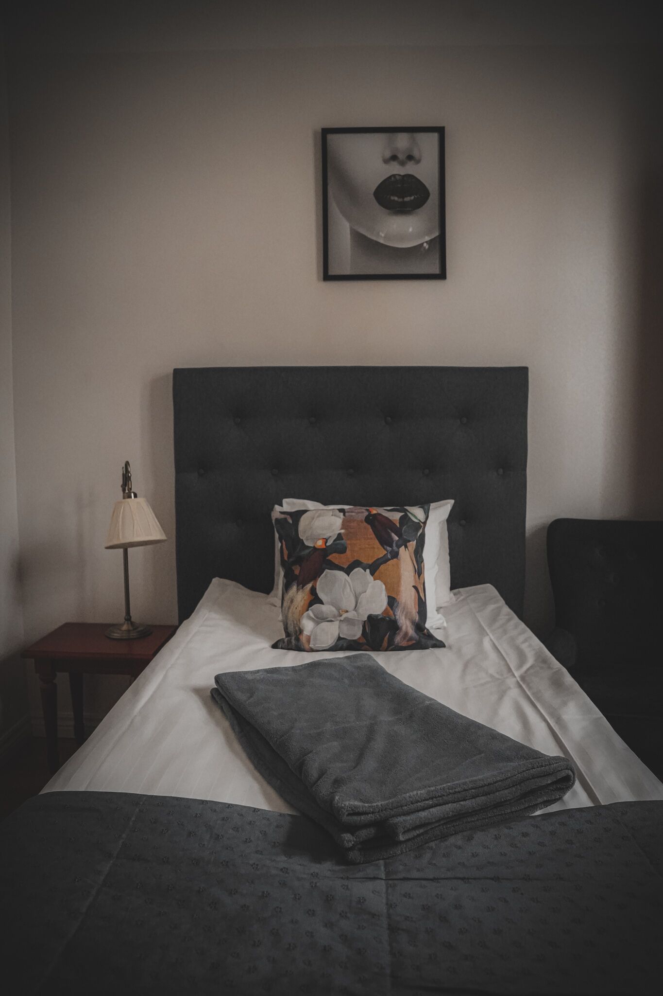 A neatly made bed with a floral pillow and a folded blanket, a bedside lamp, and a framed picture on the wall.