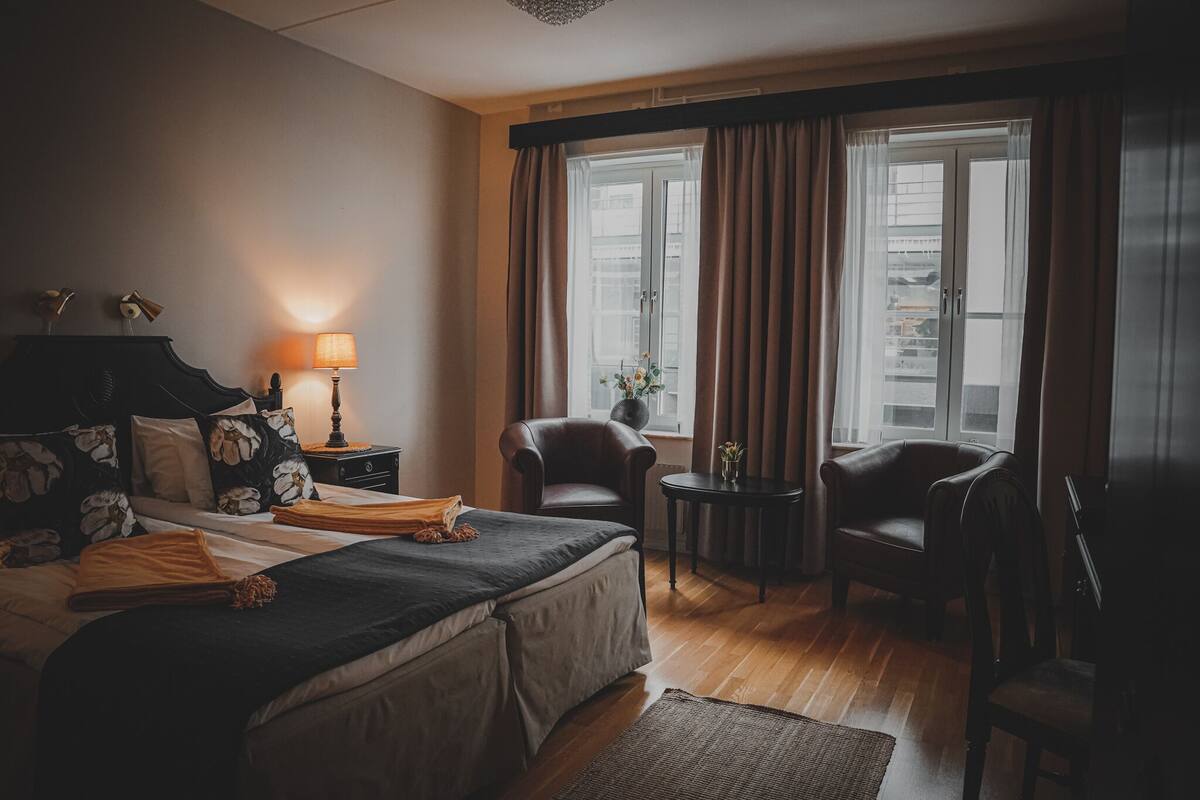 A hotel room with a bed, two chairs, a small table with a lamp, and a window with curtains.