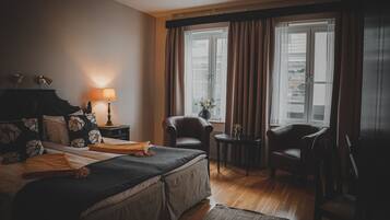 Una habitación de hotel con una cama, dos sillas, una mesita con una lámpara y un ventanal con cortinas.