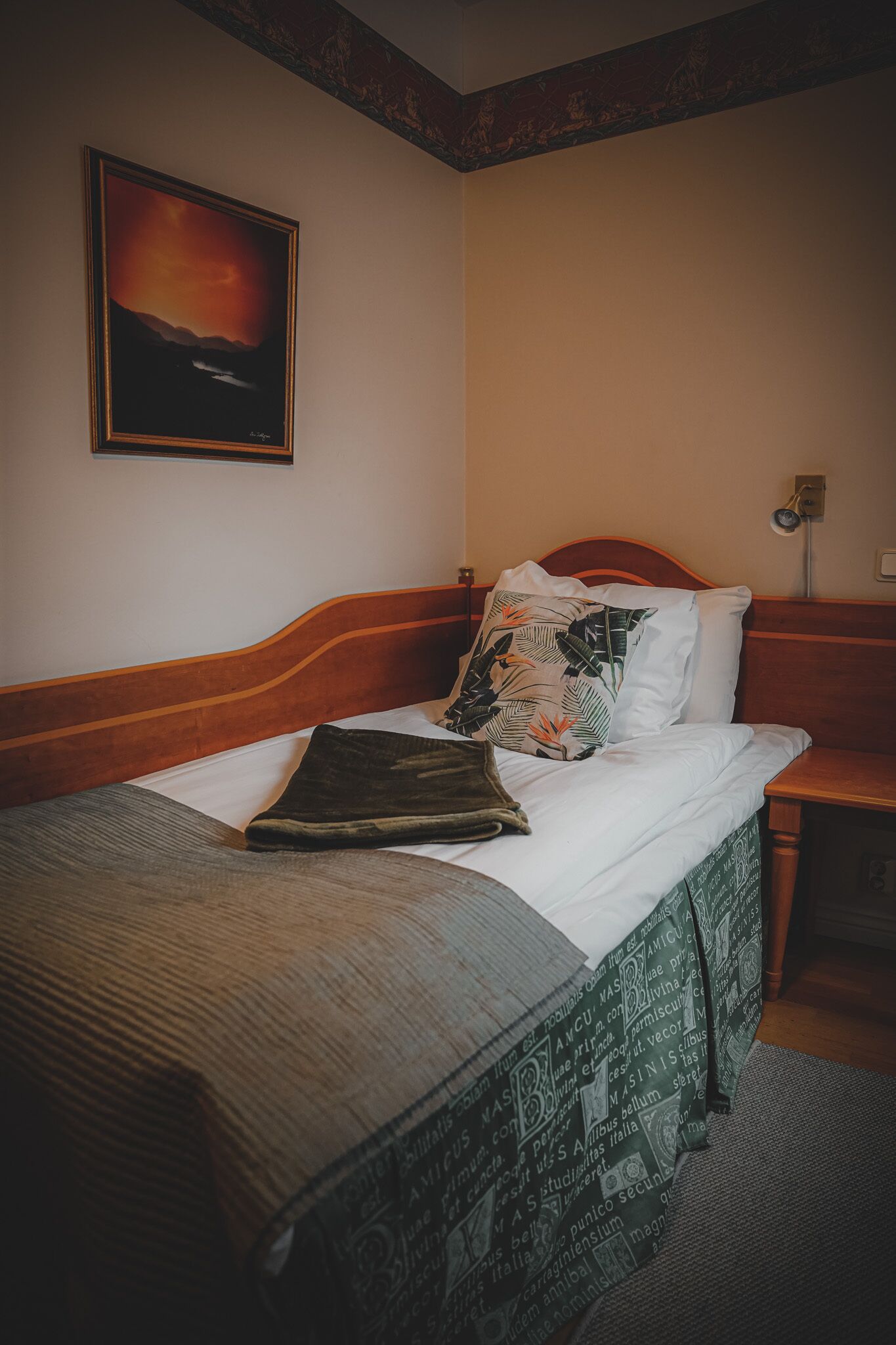 A neatly made bed with a patterned bedspread, a pillow with a tropical print, and a framed picture of a sunset on the wall.