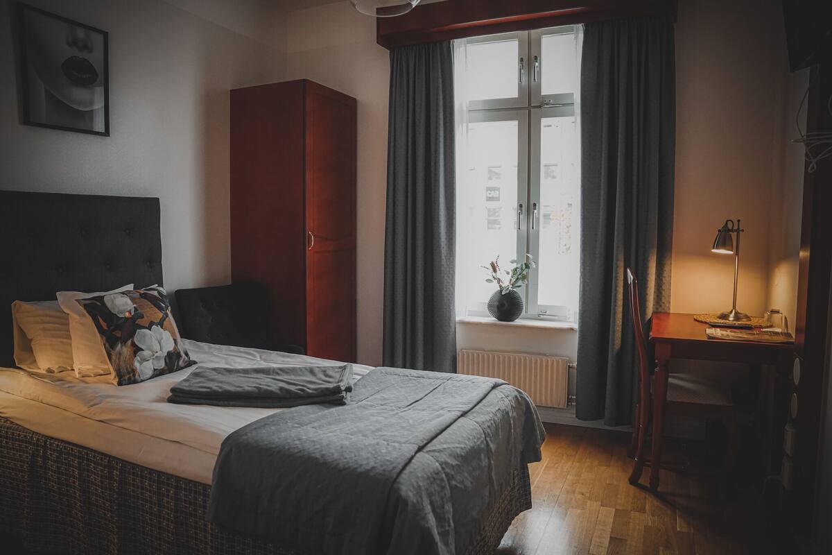 A bedroom with a bed, a wardrobe, a desk with a lamp, and a window with curtains.