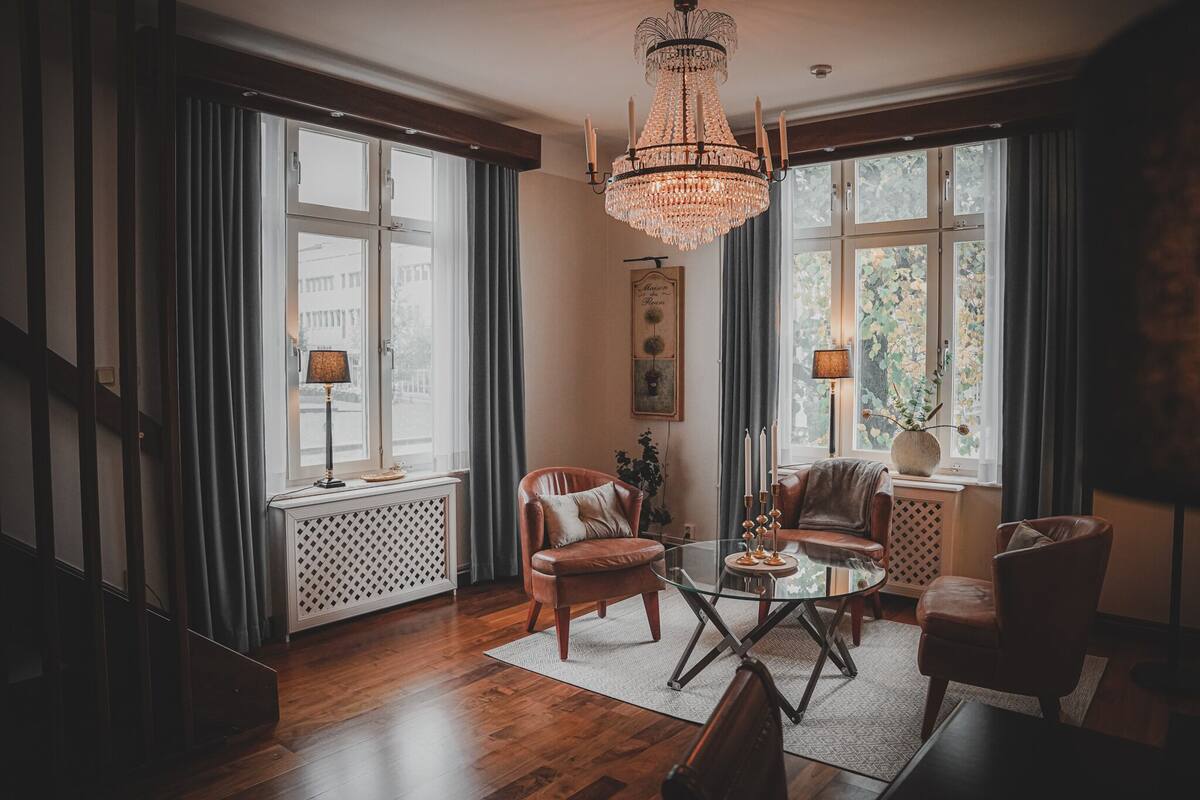 A spacious living room with a chandelier, leather chairs, a glass coffee table, and large windows with curtains.