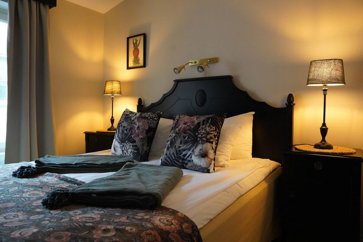 A neatly made bed with floral-patterned pillows and a dark headboard. There are two bedside lamps on either side of the bed.