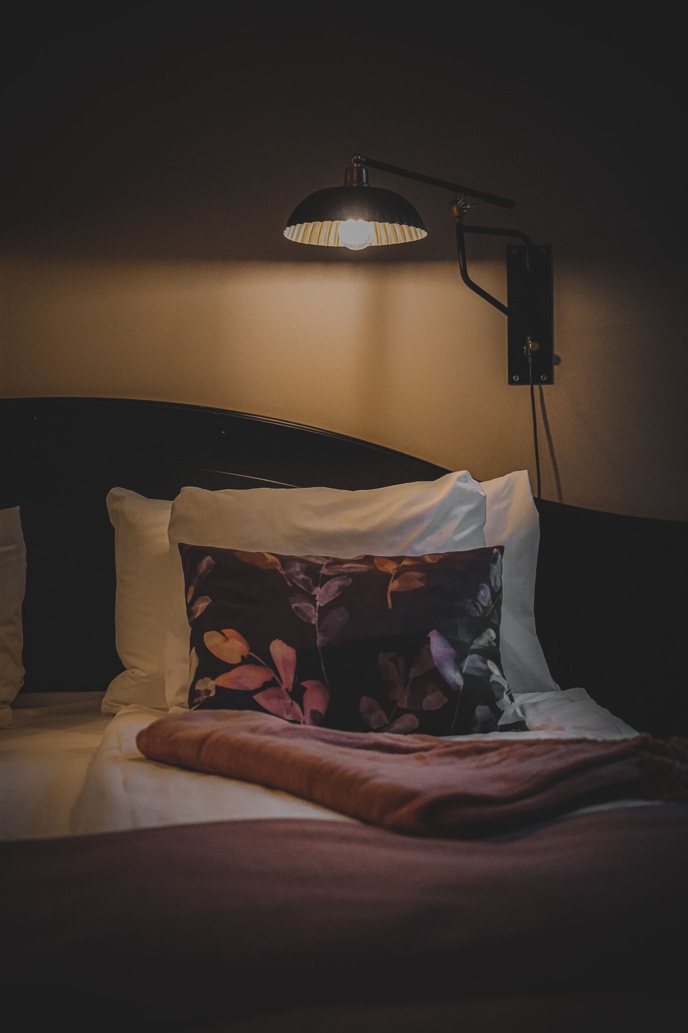 Ein Bett mit weißen und blumigen Kissen, einem dunklen Kopfteil und einer einzelnen Nachttischlampe.