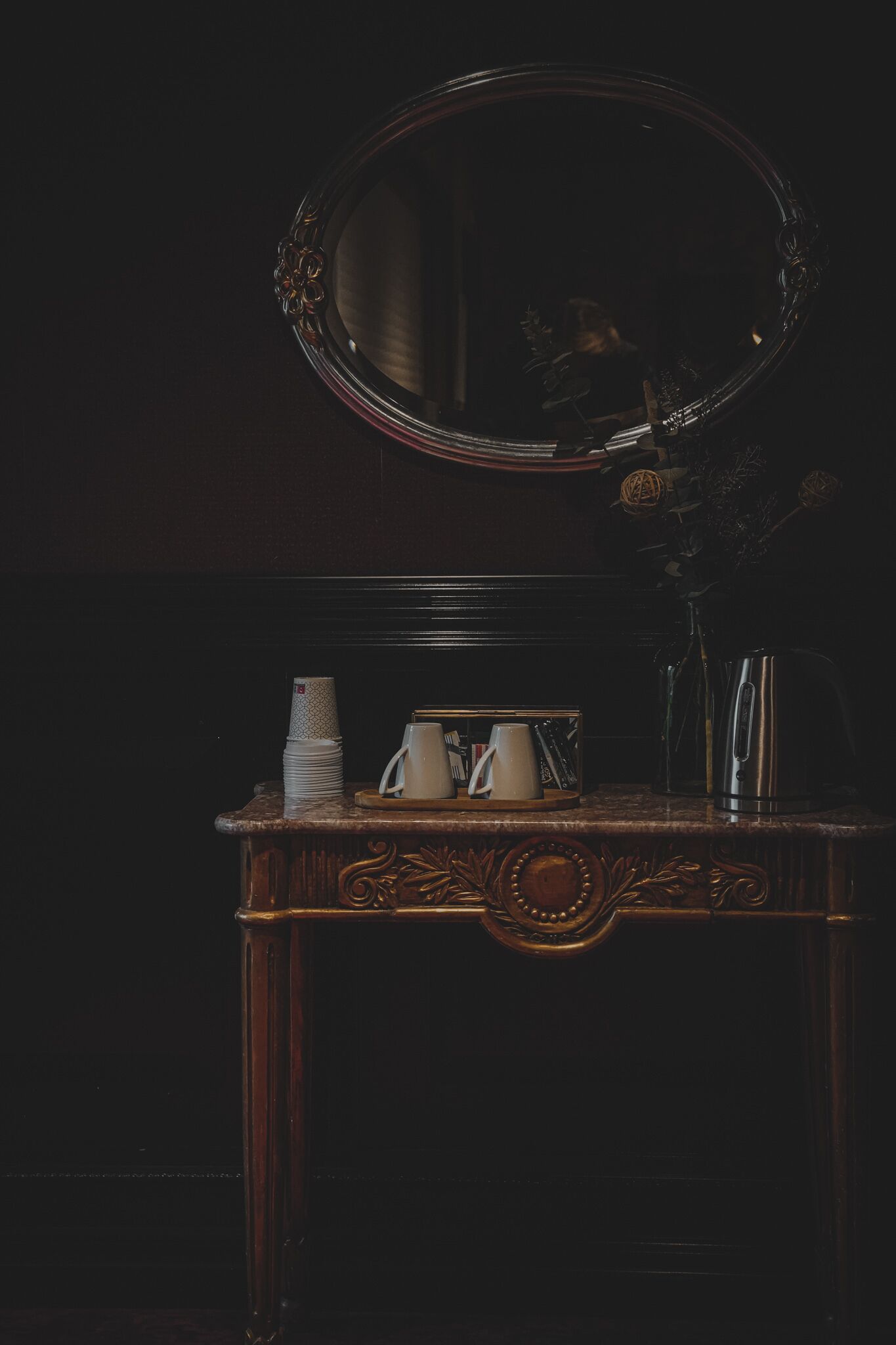 A dark wooden vanity with a round mirror, a stack of white cups, a white pitcher, and a kettle.