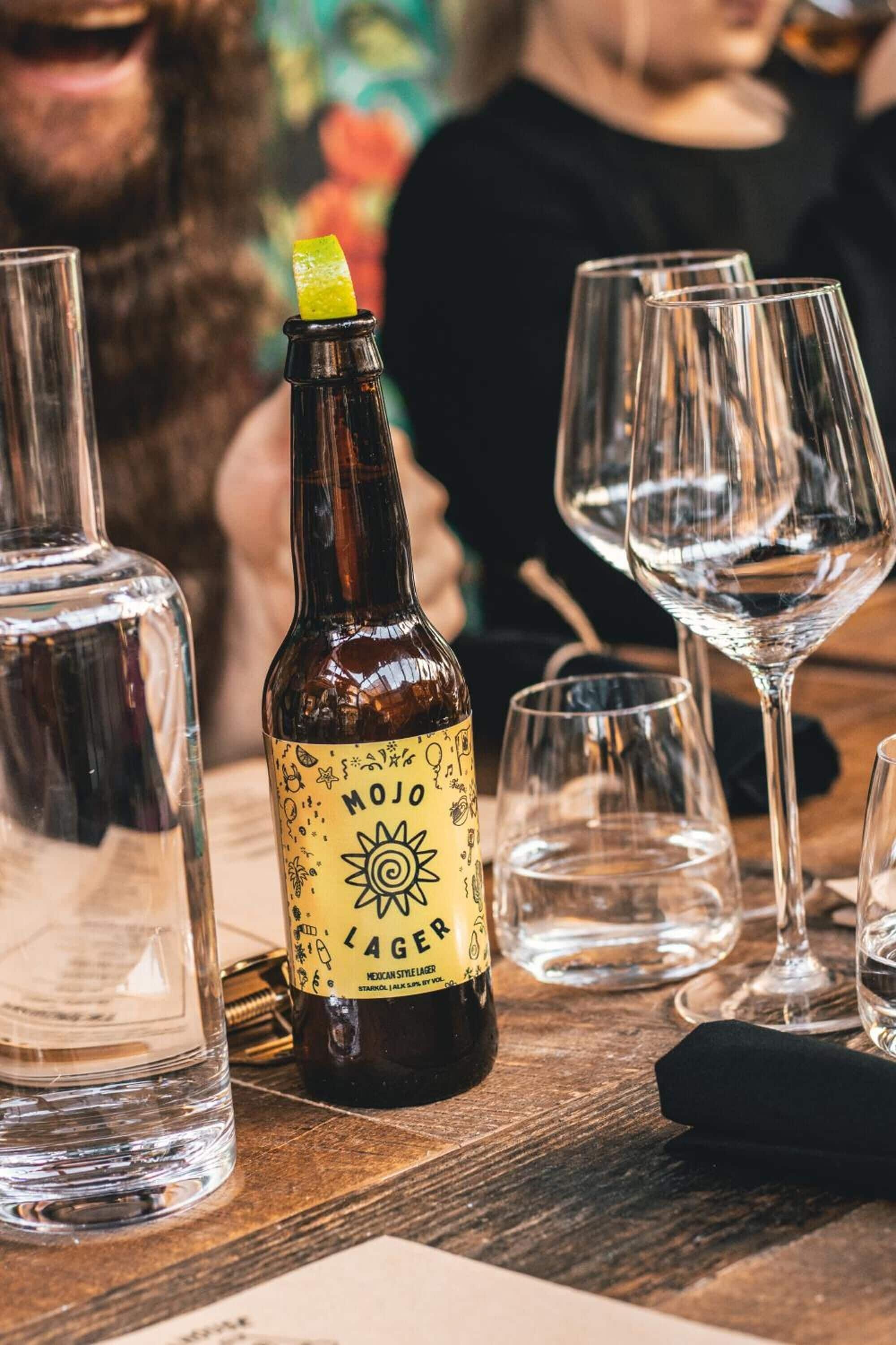 Una botella de cerveza Mojo Lager con etiqueta que muestra un sol y varios iconos, rodeada de vasos vacíos sobre una mesa de madera.