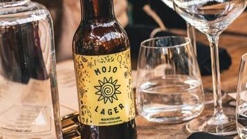Una botella de cerveza Mojo Lager con etiqueta que muestra un sol y varios iconos, rodeada de vasos vacíos sobre una mesa de madera.